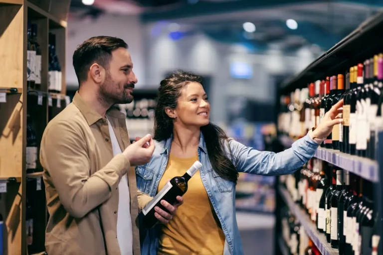 Couple shopping for wine: woman choosing a bottle, man holding wine in hand at supermarket shelf