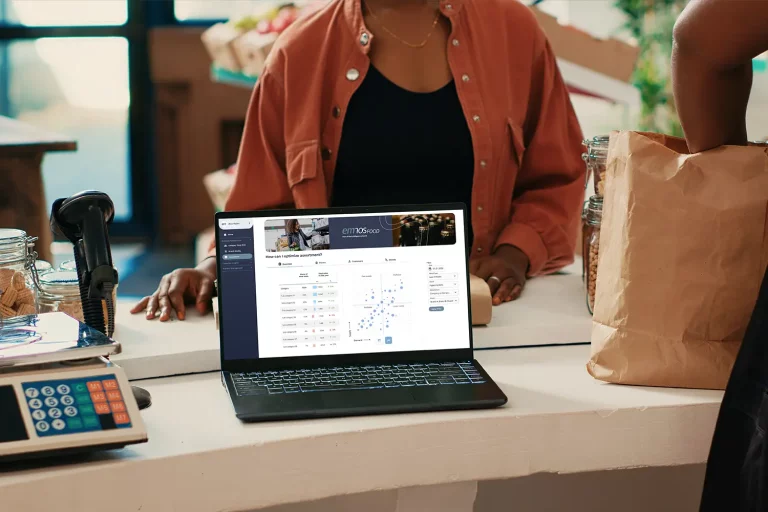 Retail employee standing at checkout counter with laptop displaying emnosFOOD retail analytics platform interface for assortment optimization.
